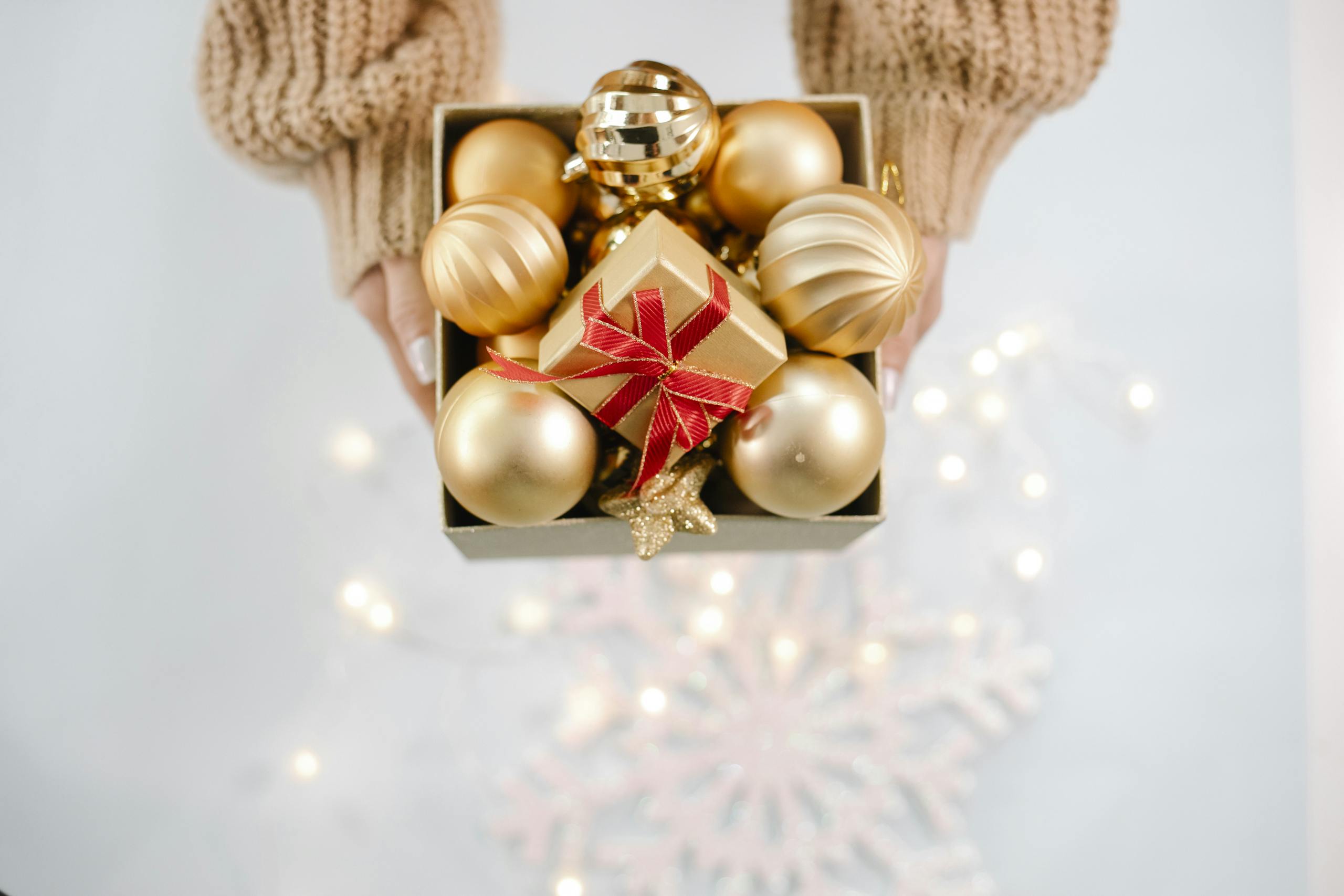 Prestations à la carte Décembre From above of crop anonymous woman in sweater showing box with golden Christmas baubles against light background with glowing garland
